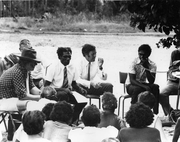 Middle range shot of a group 16 people seated in chairs in a circle in the bush. Six people are facing the camera, the rest have their backs to the camera.